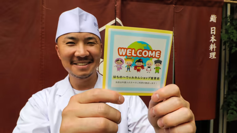 Daisuke Tanaka, quản lý của một nhà hàng Hachinohe có tên là Botejuu, giơ một nhãn dán để báo hiệu rằng khách du lịch được chào đón. (Ảnh của Ken Kobayashi)
